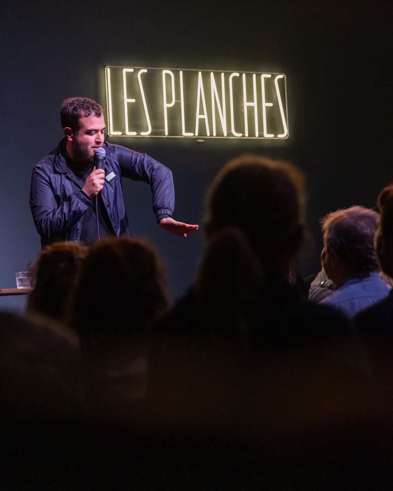 Un humoriste en stand-up devant le public, sous le néon Les Planches