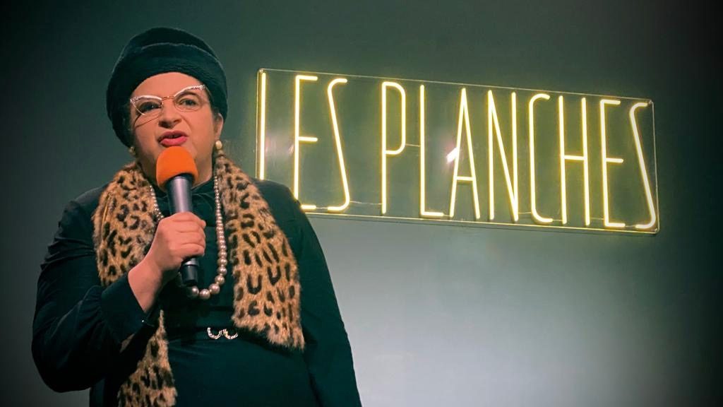 Une humoriste en personnage, micro orange, devant le néon